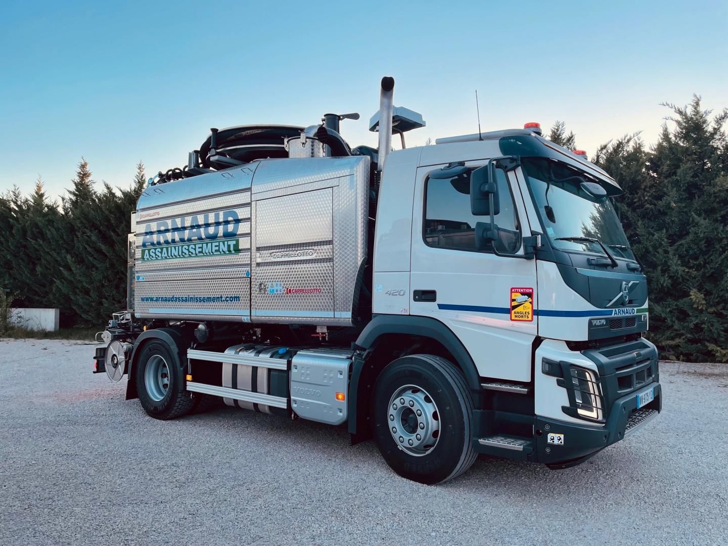 Camion de ramassage des ordures blanc et argent sur fond de gravier, ciel bleu. Le logo de l'entreprise est apposé sur le côté du camion.