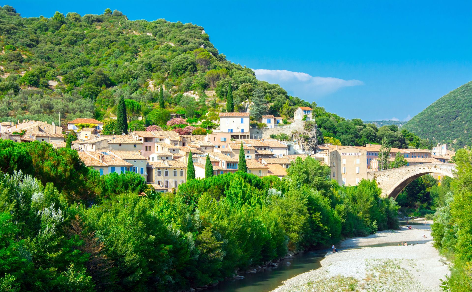 Un village aux bâtiments en pierre et au pont arqué, niché au bord d'une rivière et de collines verdoyantes sous un ciel bleu.