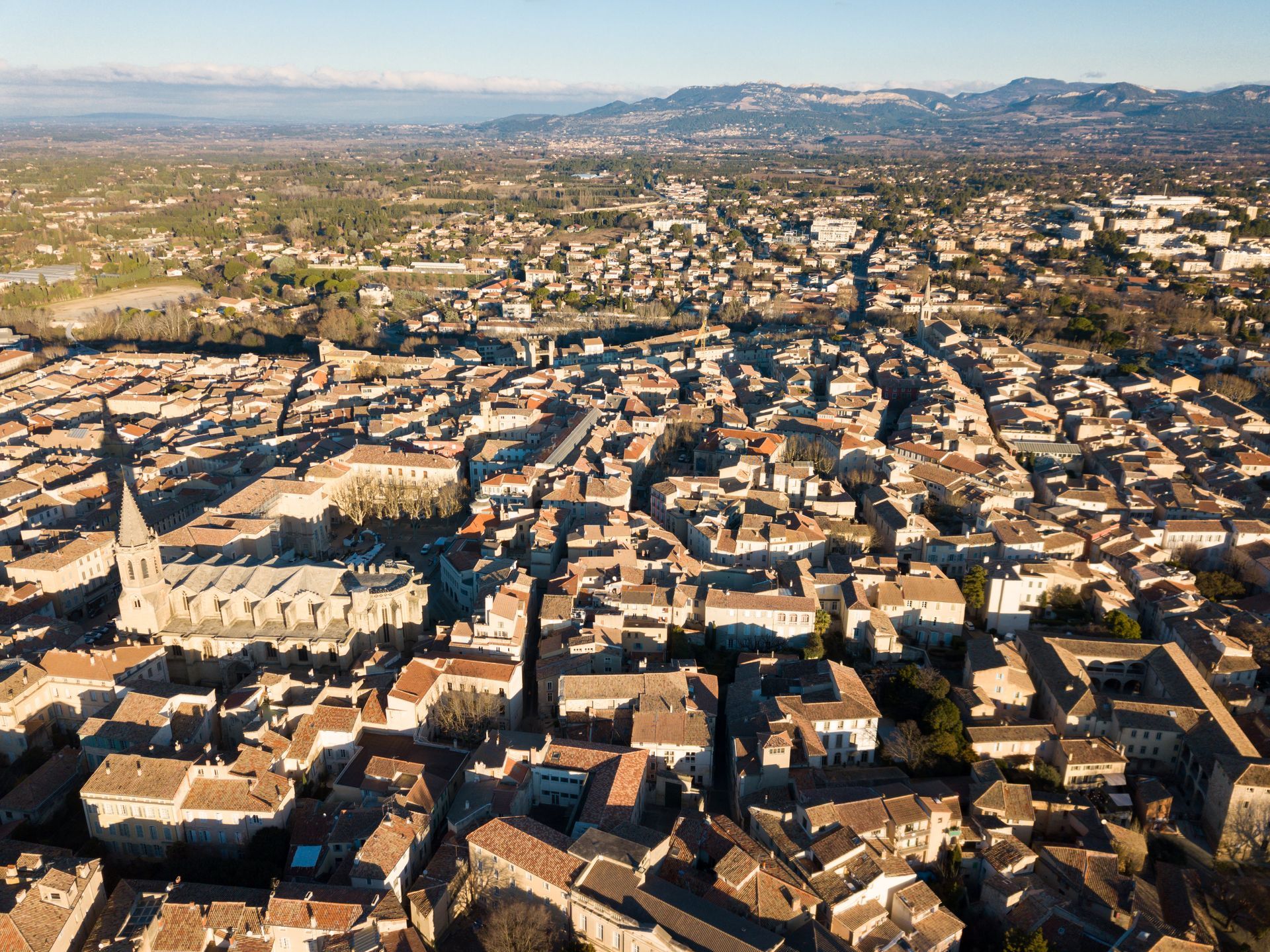 Vue aérienne d'une ville européenne aux toits de tuiles rouges, aux bâtiments et à la cathédrale. Montagnes brumeuses à l'horizon.