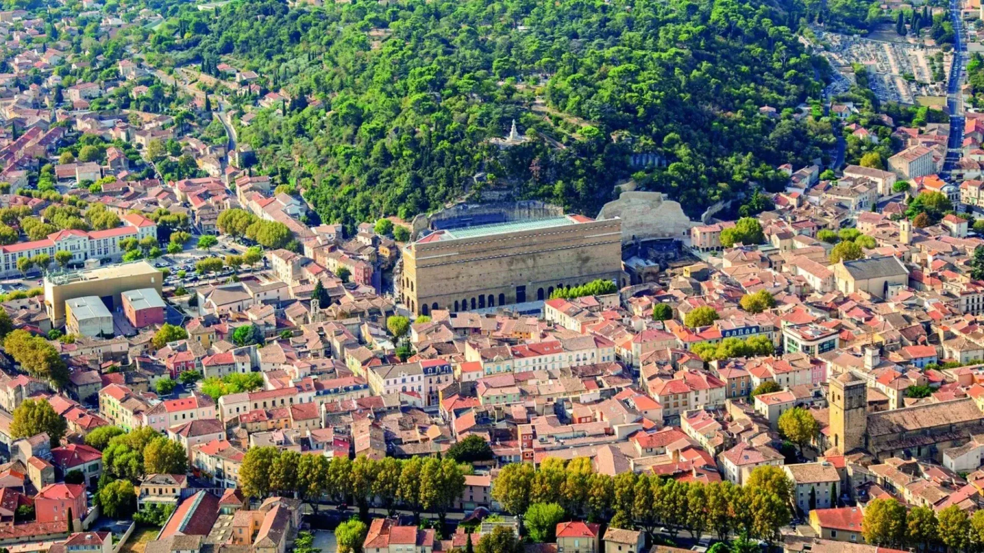 Vue aérienne d'une ville aux toits de tuiles rouges, d'un grand théâtre romain et d'une colline verdoyante et boisée.