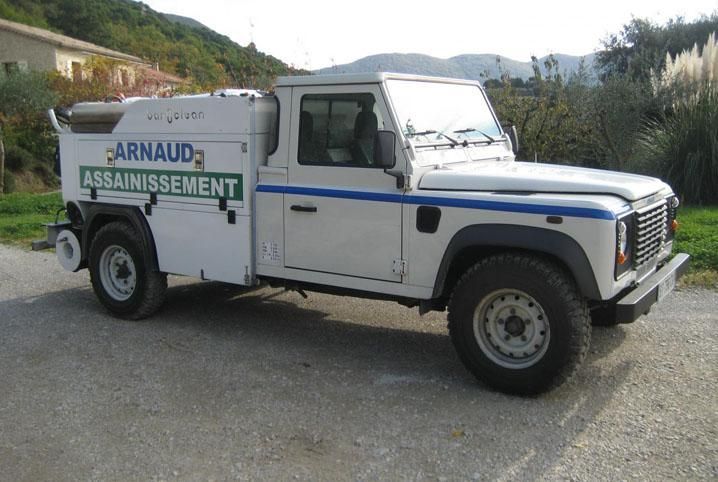 Camion de travail Land Rover Defender blanc avec bande bleue et inscription « ARNAUD Assainissement » sur le côté.