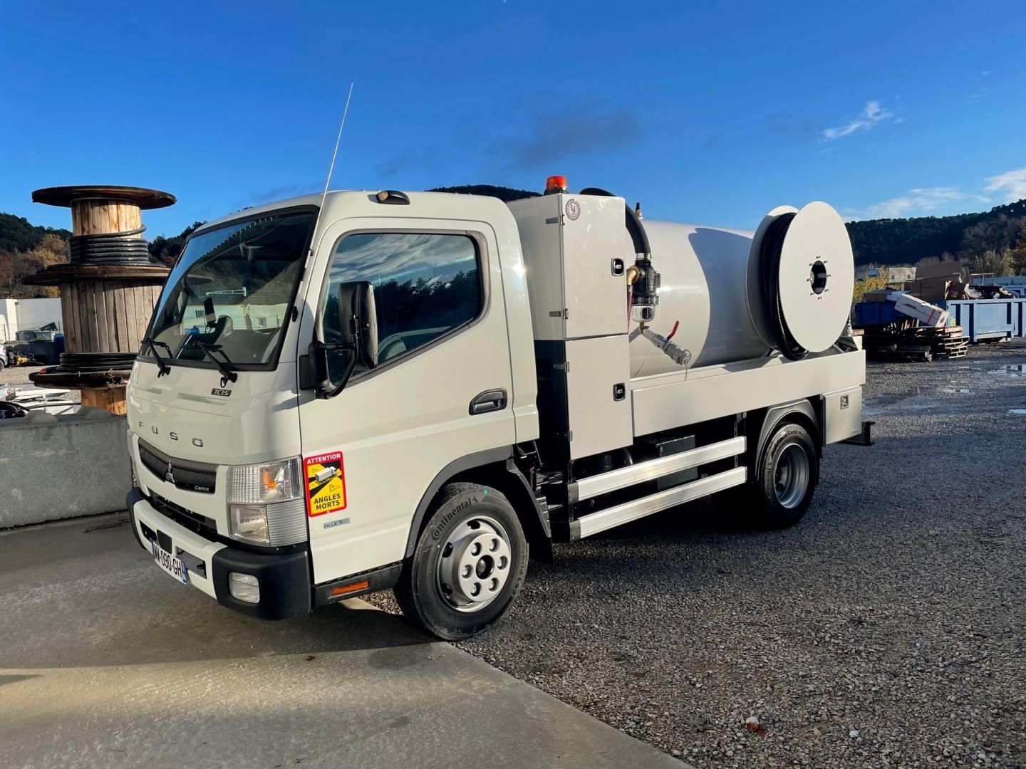 Camion de travail blanc avec réservoir et bobine, en extérieur par une journée ensoleillée.