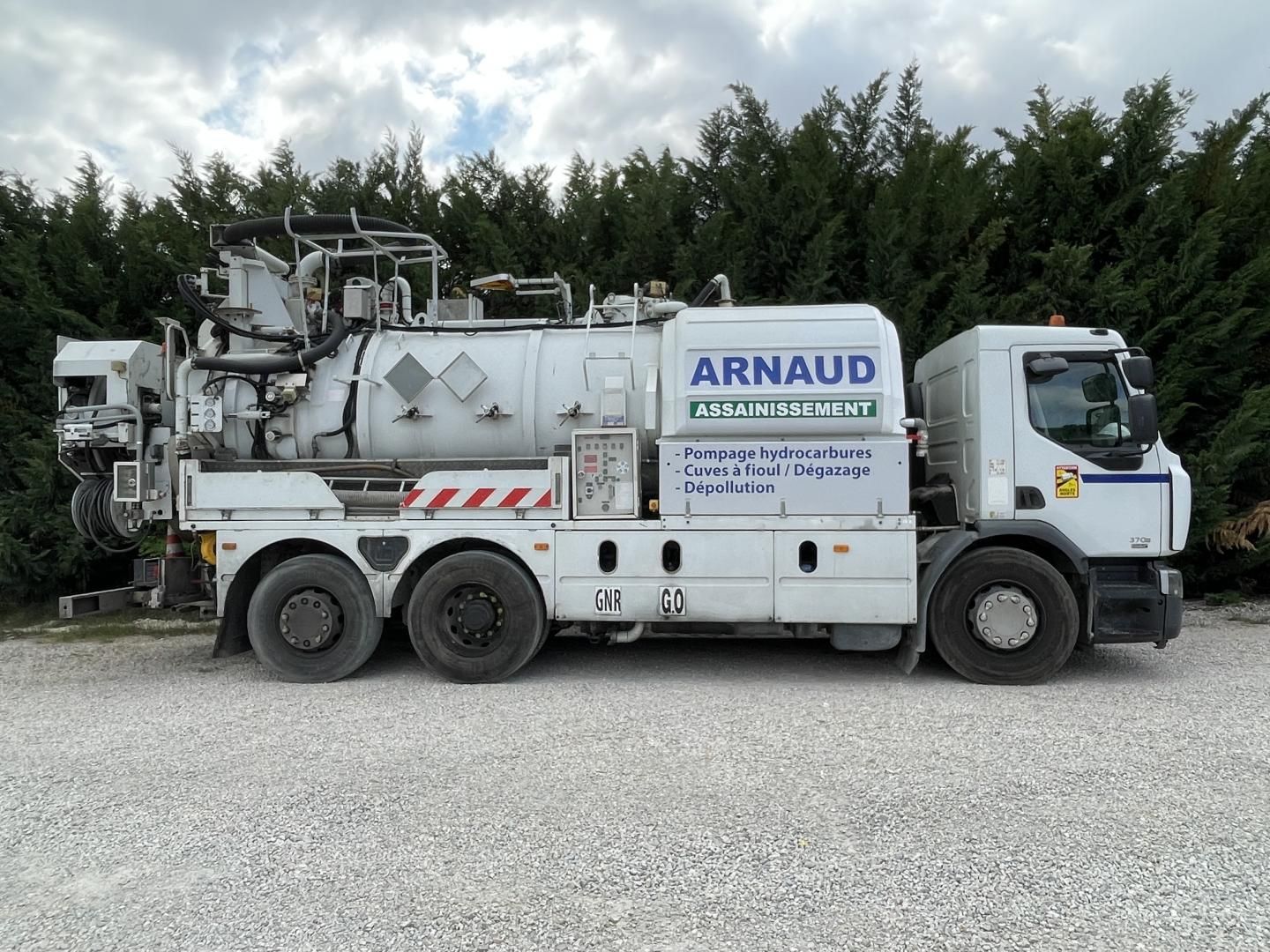 Camion industriel blanc Arnaud sur un terrain gravillonné ; arrière-plan d'arbres et de ciel nuageux.
