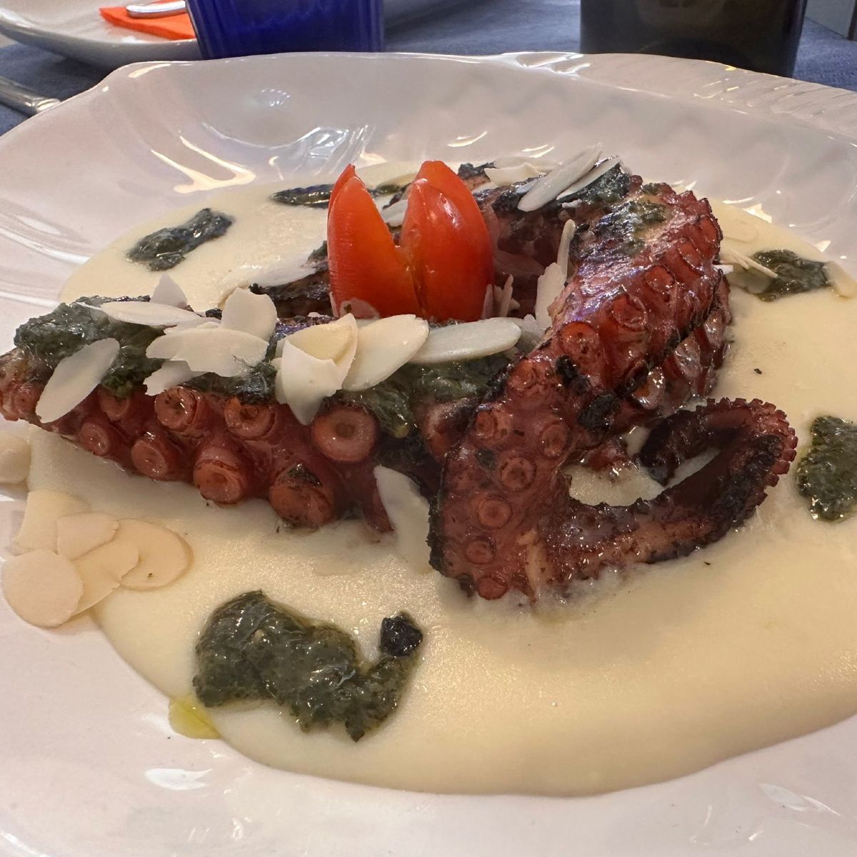 Pulpo a la parrilla con salsa blanca, pesto, almendras en hojuelas y tomates cherry en un plato blanco.