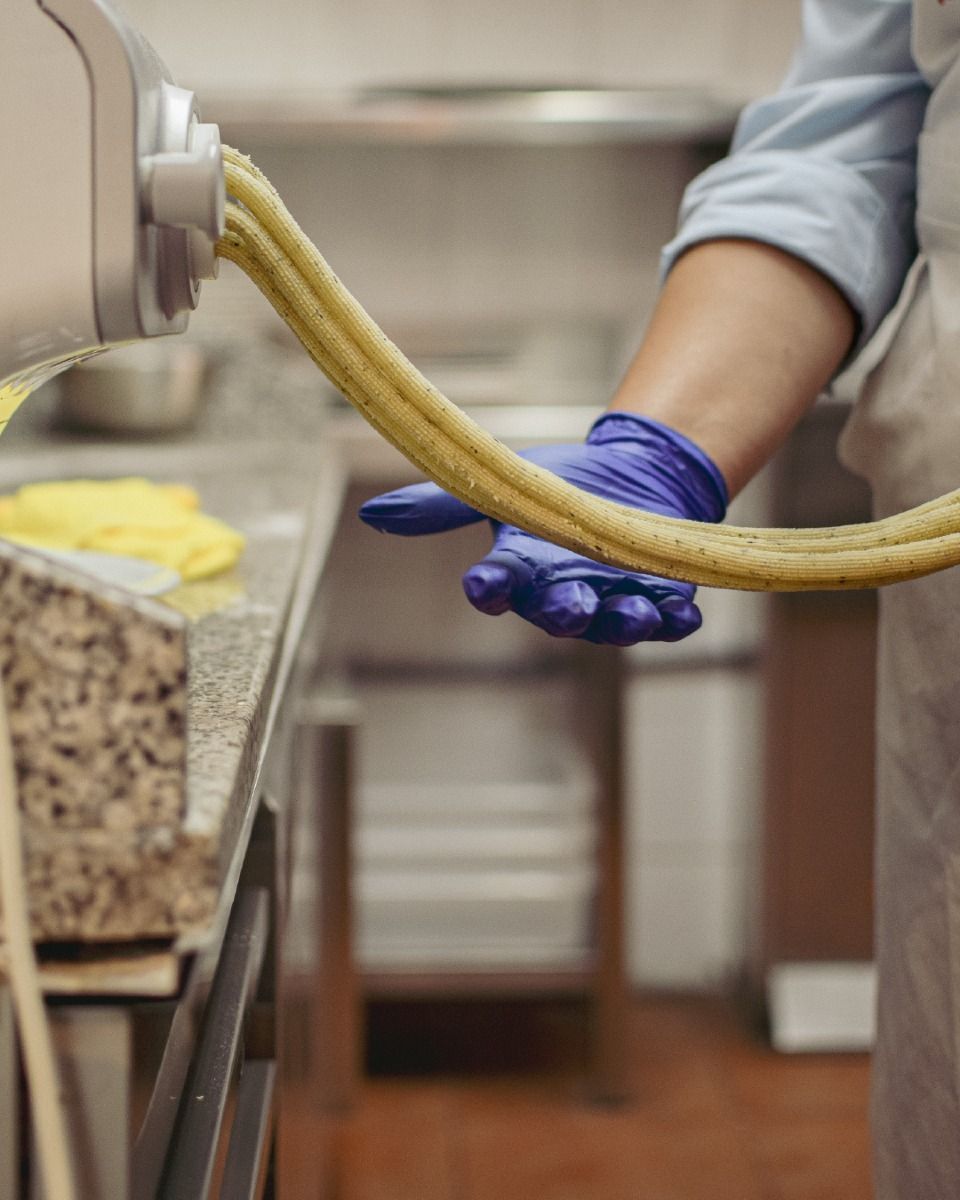 Una persona con guantes azules y un delantal blanco saca pasta de una máquina en una cocina.