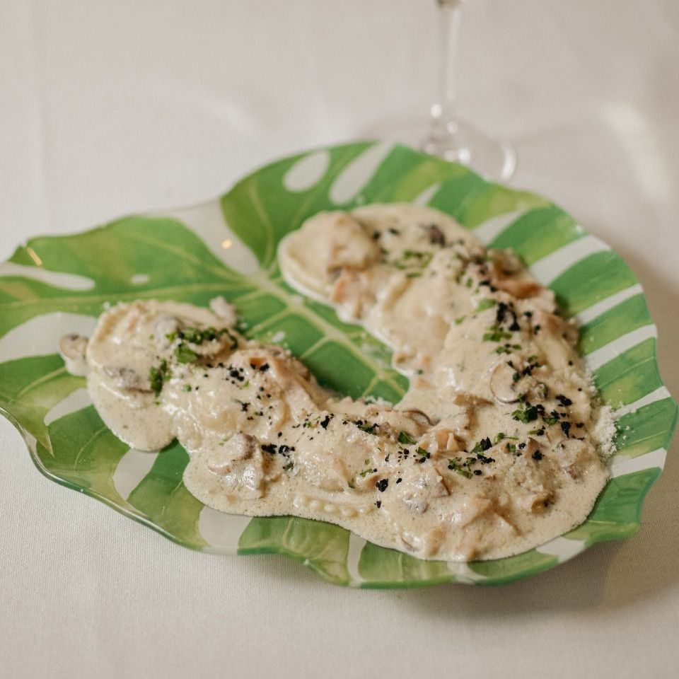 Raviolis en salsa cremosa con champiñones sobre un plato verde y blanco con forma de hoja; una copa de vino en el fondo.