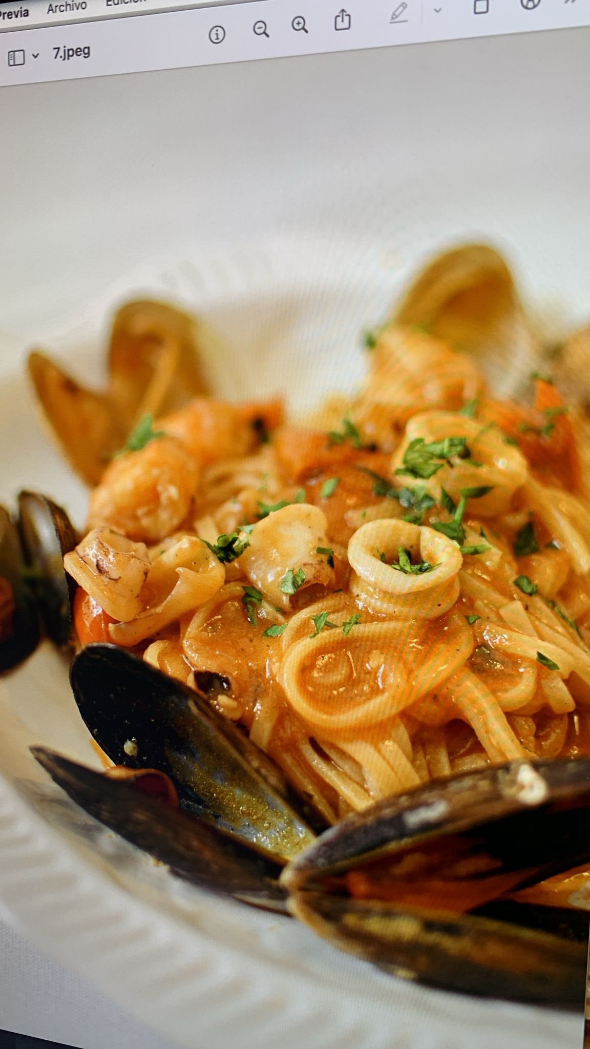 Plato de pasta con mariscos: camarones, mejillones y calamares en salsa roja, adornados con perejil en un plato blanco.