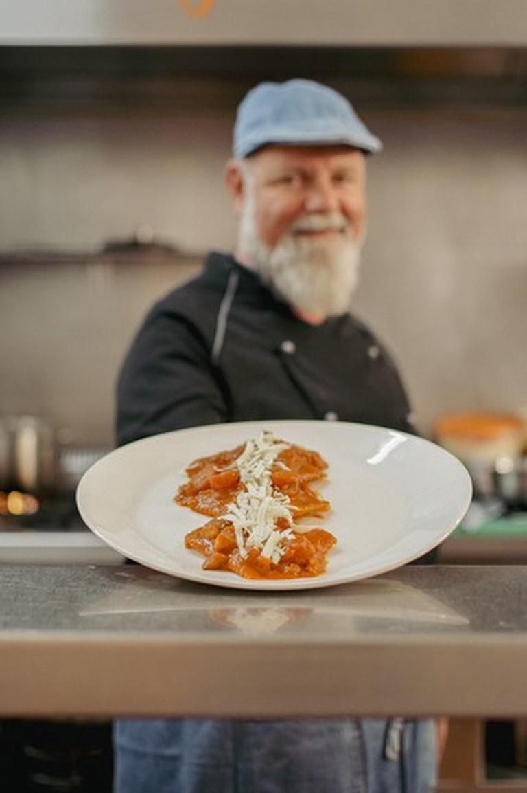 Chef con una gorra azul, sosteniendo un plato de comida con una sonrisa, en un entorno de cocina.
