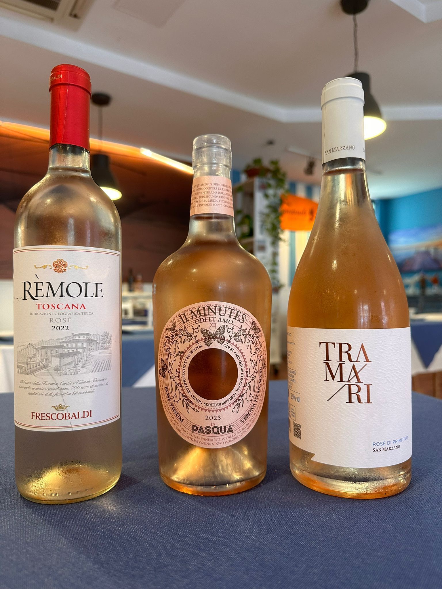 Tres botellas de vino de diferentes colores, sobre un mantel azul, con un ambiente de restaurante al fondo.