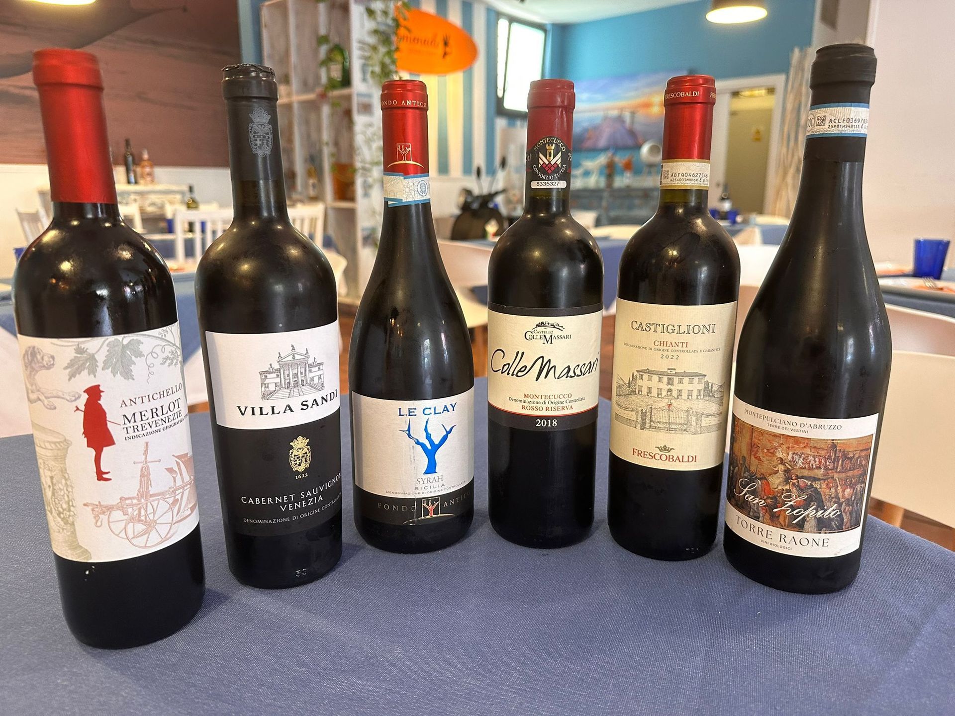 Seis botellas de vino tinto en una mesa, cada una con una etiqueta diferente, en un entorno de restaurante.