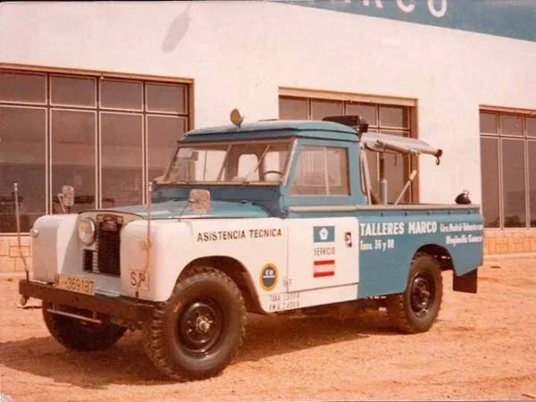 Grúa antigua azul y blanca estacionada frente a un edificio en un terreno de tierra.