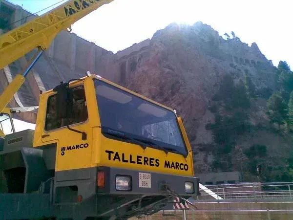 Un camión grúa amarillo con la inscripción “TALLERES MARCO” estacionado en una obra de construcción en la montaña rocosa.