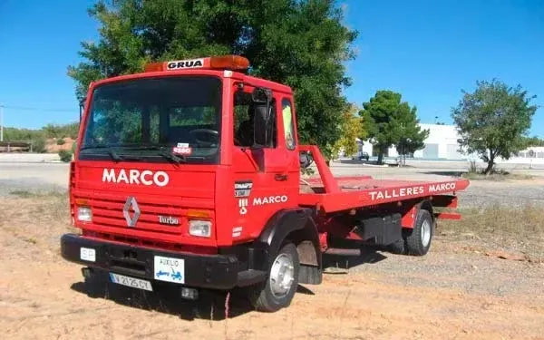 Grúa roja con plataforma estacionada al aire libre en un terreno de tierra bajo unos árboles.