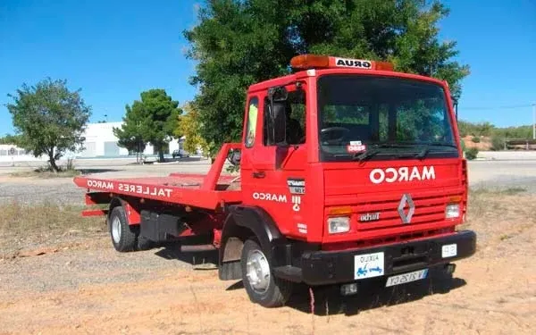 Grúa roja con plataforma estacionada al aire libre en un terreno de tierra bajo unos árboles.