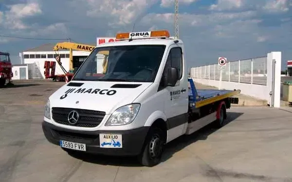 Una grúa blanca de la marca Marco está estacionada al aire libre, con la plataforma ligeramente elevada, frente a un edificio.
