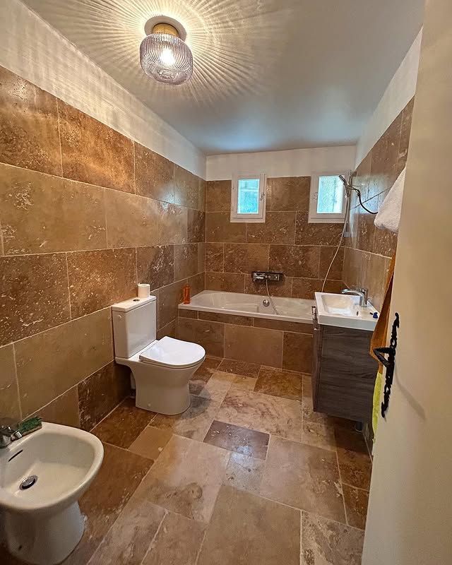 Salle de bains aux murs carrelés de brun, baignoire, toilettes, bidet et petites fenêtres. Un luminaire est fixé au plafond.