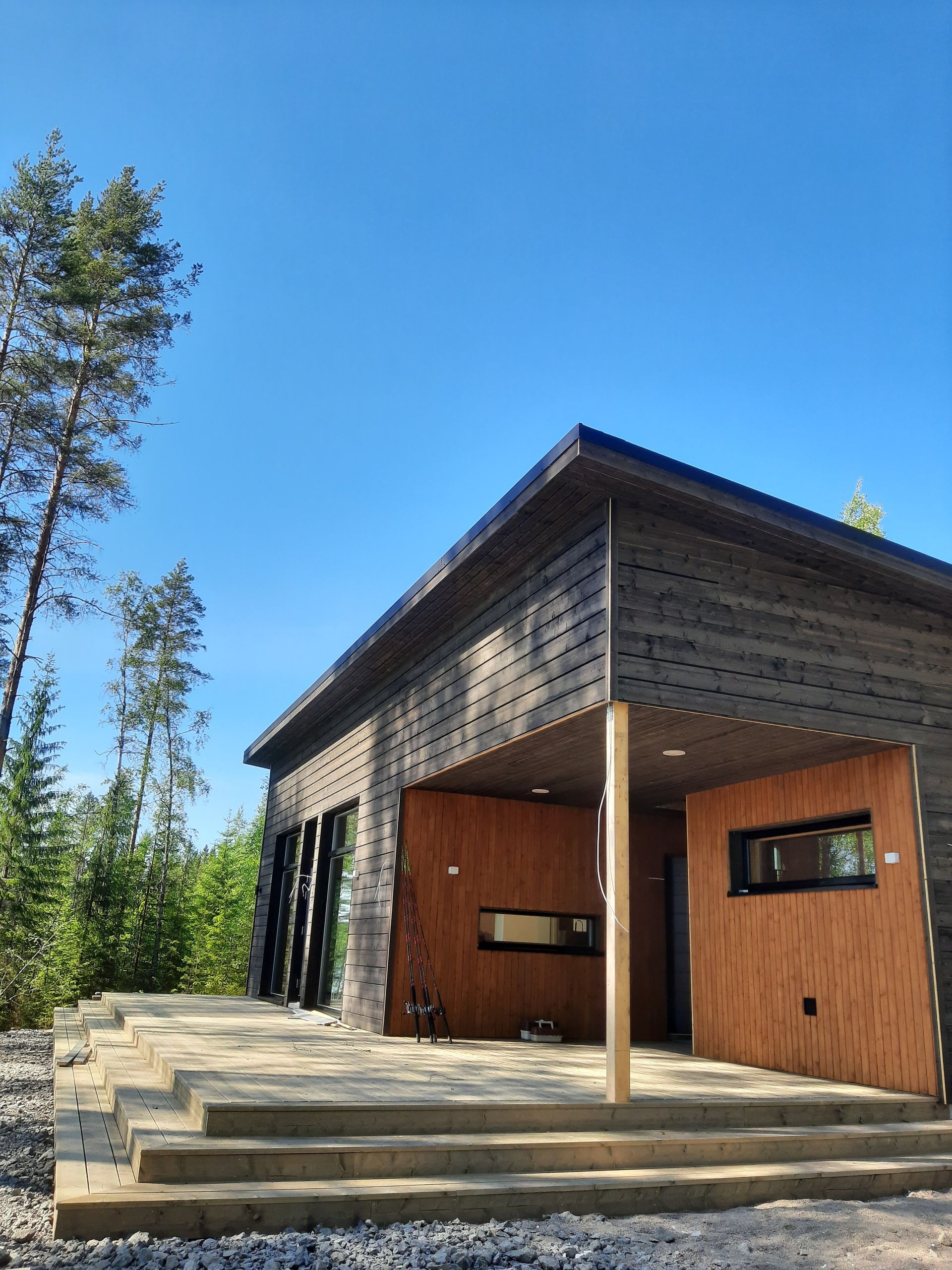 Moderni puinen mökki, jossa on suuret ikkunat ja porrastettu kivinen patio sinisen taivaan alla