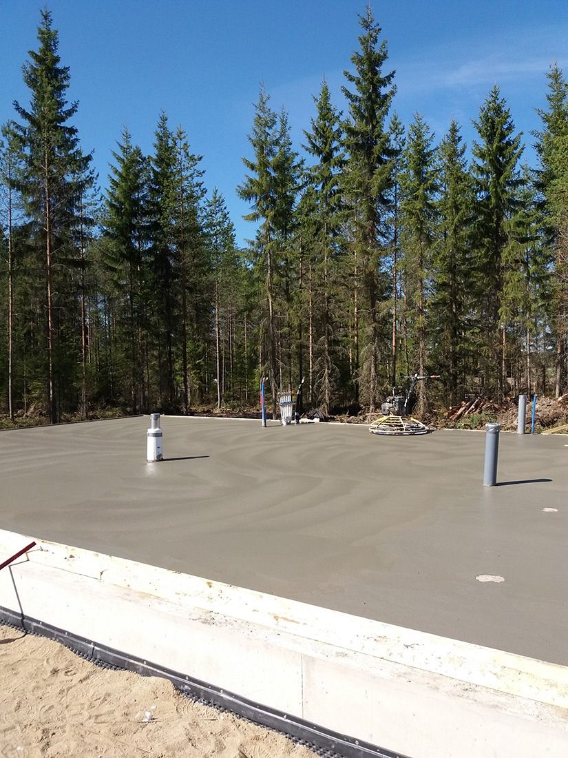 Betoninen perustuslaatta metsäisellä aukiolla, jossa näkyvissä putkia ja sähköpylväitä sinisen taivaan alla