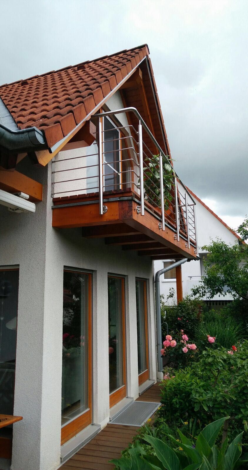 Zweistöckiges Haus mit rotem Ziegeldach, weißer Fassade, Balkon und Gartenpflanzen am Eingang
