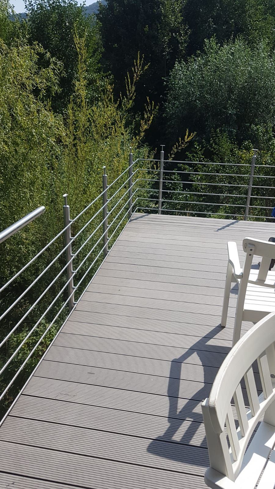 Terrasse mit Metallgeländer neben dichtem Grün; rechts werfen weiße Stühle Schatten.