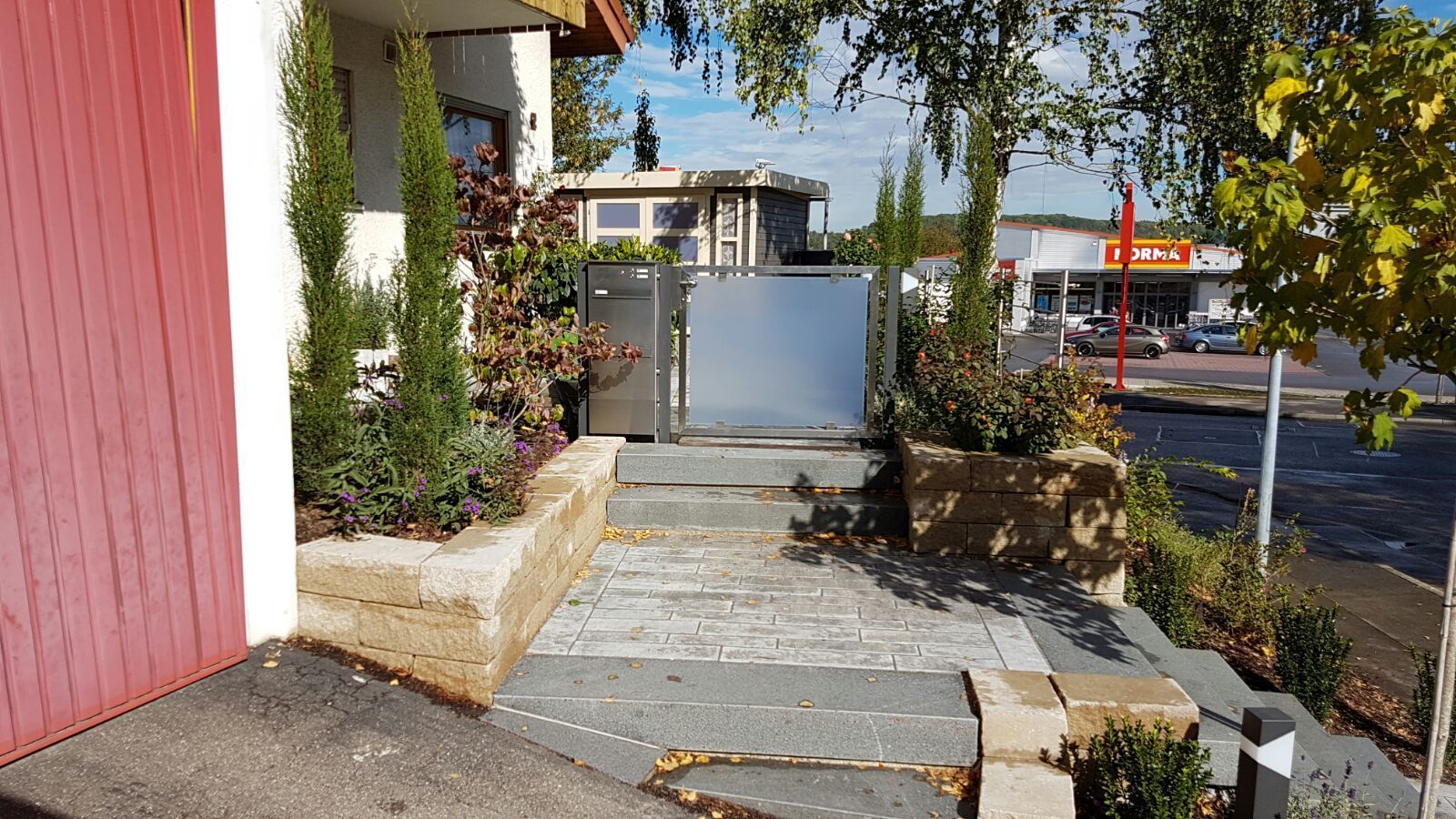 Ein Steinweg neben einem rosafarbenen Gebäude führt zu einem Tor und einer Straße mit Pflanzkästen und Sträuchern.