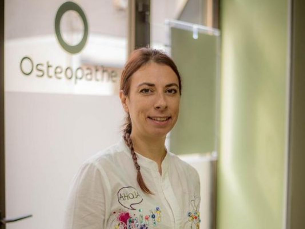 Une femme en uniforme blanc se tient dans l'embrasure de la porte d'une clinique d'ostéopathie, une pancarte se trouvant derrière elle.