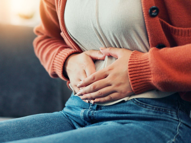 Une personne se tient le bas du ventre en étant assise, vêtue d'un jean et d'un cardigan orange.