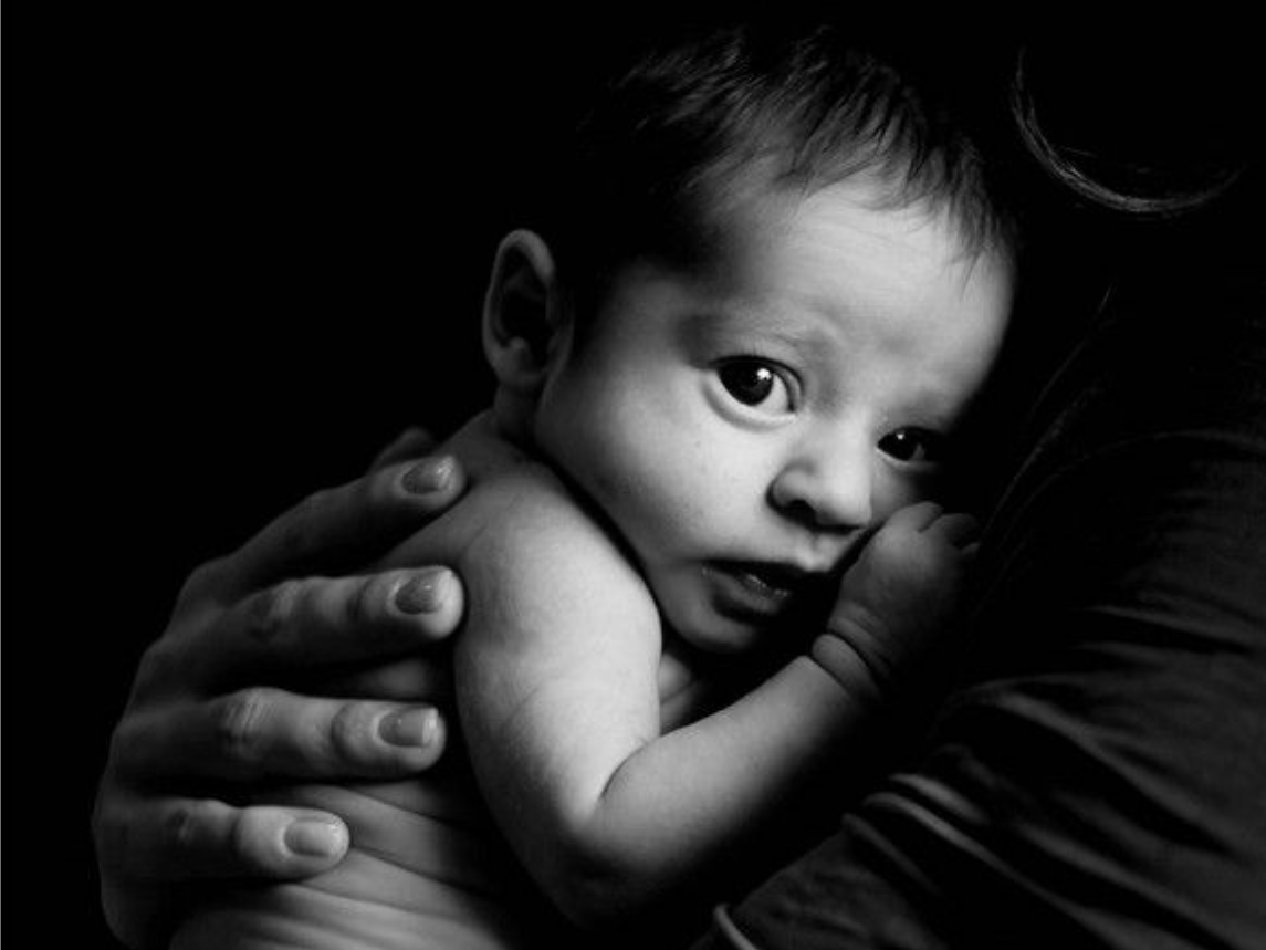 Un bébé blotti dans les bras d'un adulte, regardant l'objectif dans une douce lumière en noir et blanc.