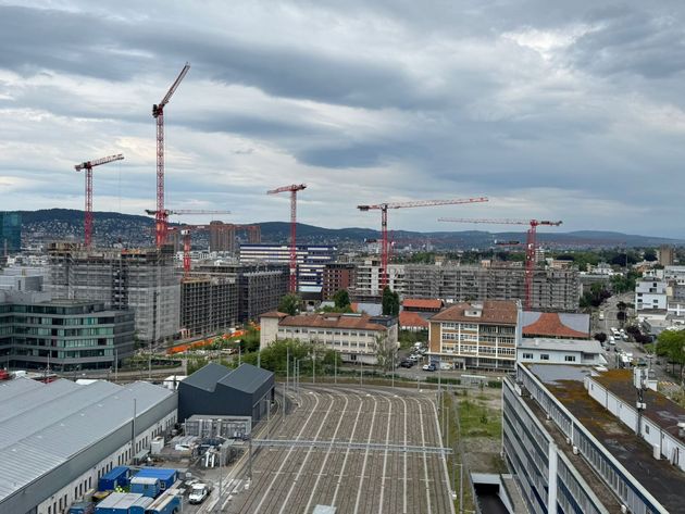 Weite Ansicht einer städtischen Baustelle mit mehreren roten Kränen, die sich unter einem bewölkten Himmel über die Gebäude erheben.