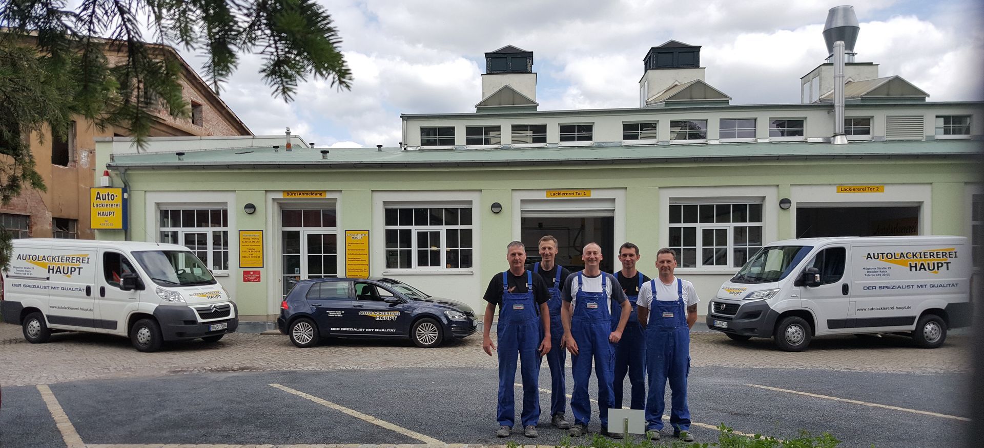Fünf Personen in blauen Overalls stehen vor einem Gebäude, an dessen Seiten zwei weiße Lieferwagen geparkt sind.