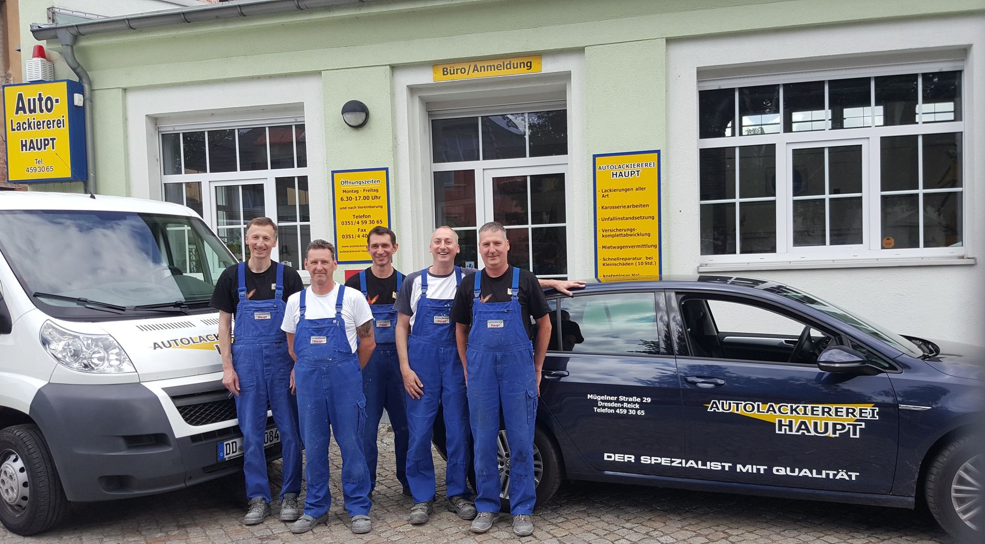 Vier Personen in blauen Overalls stehen vor einer Autowerkstatt neben Lieferwagen und einem Auto.