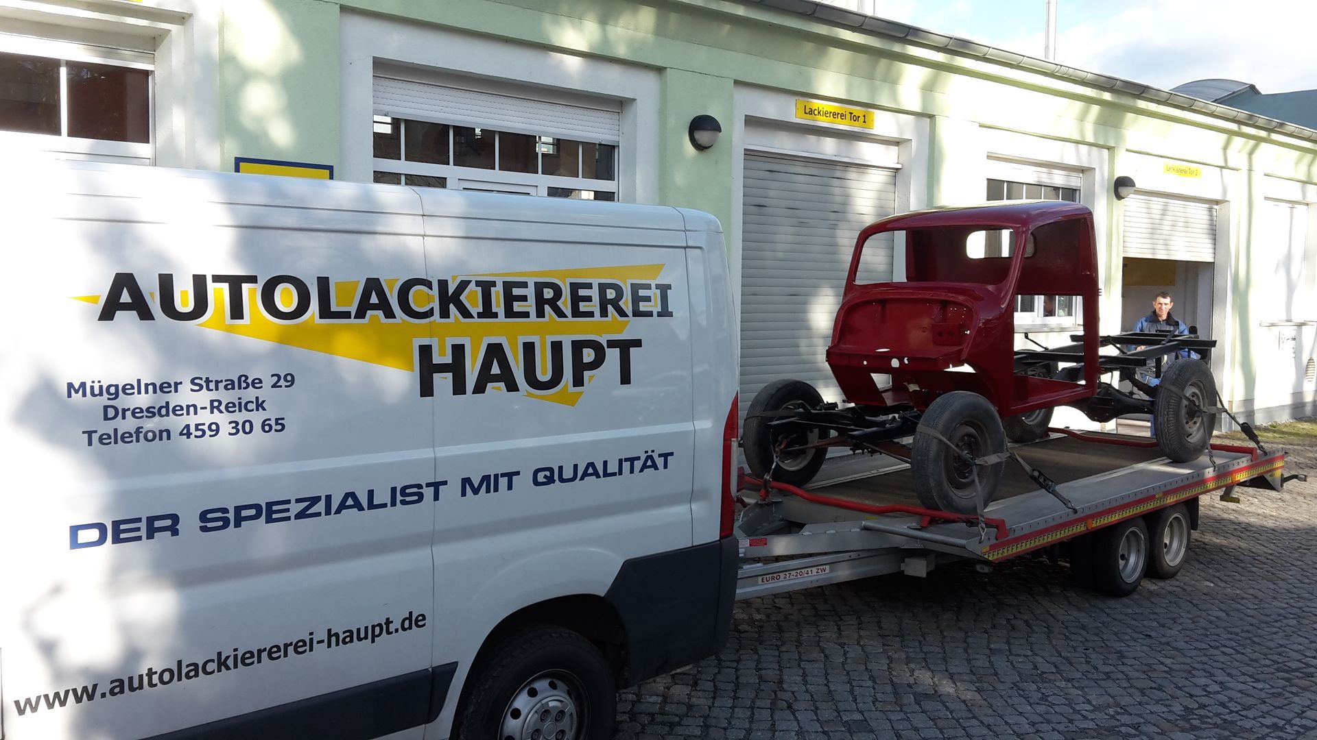Rote Autokarosserie auf einem Tieflader neben einem Lieferwagen mit der Aufschrift „Autolackiererei Haupt“.