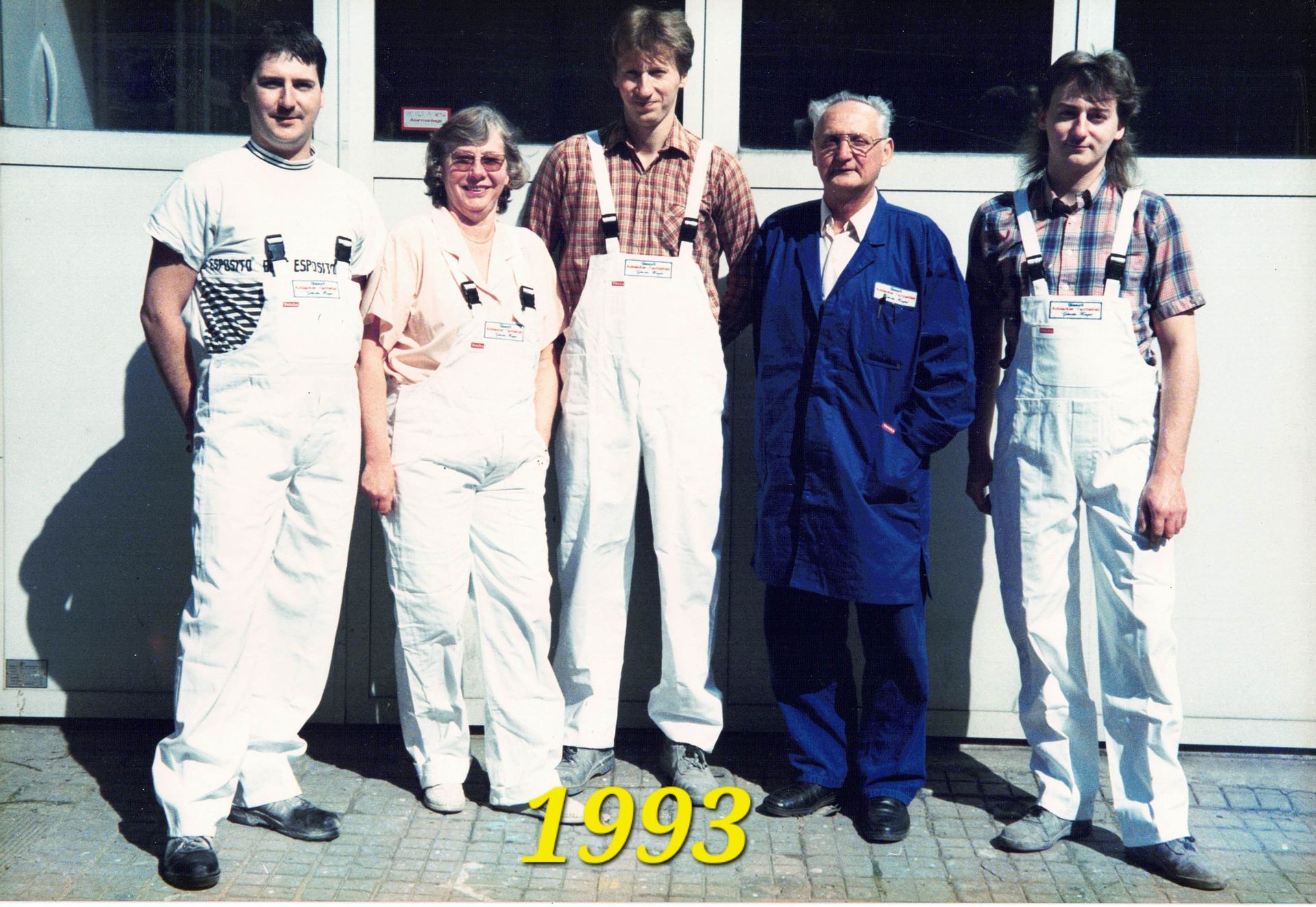 Fünf Personen stehen im Freien vor einem Gebäude, am unteren Rand ist die Jahreszahl „1993“ zu sehen.