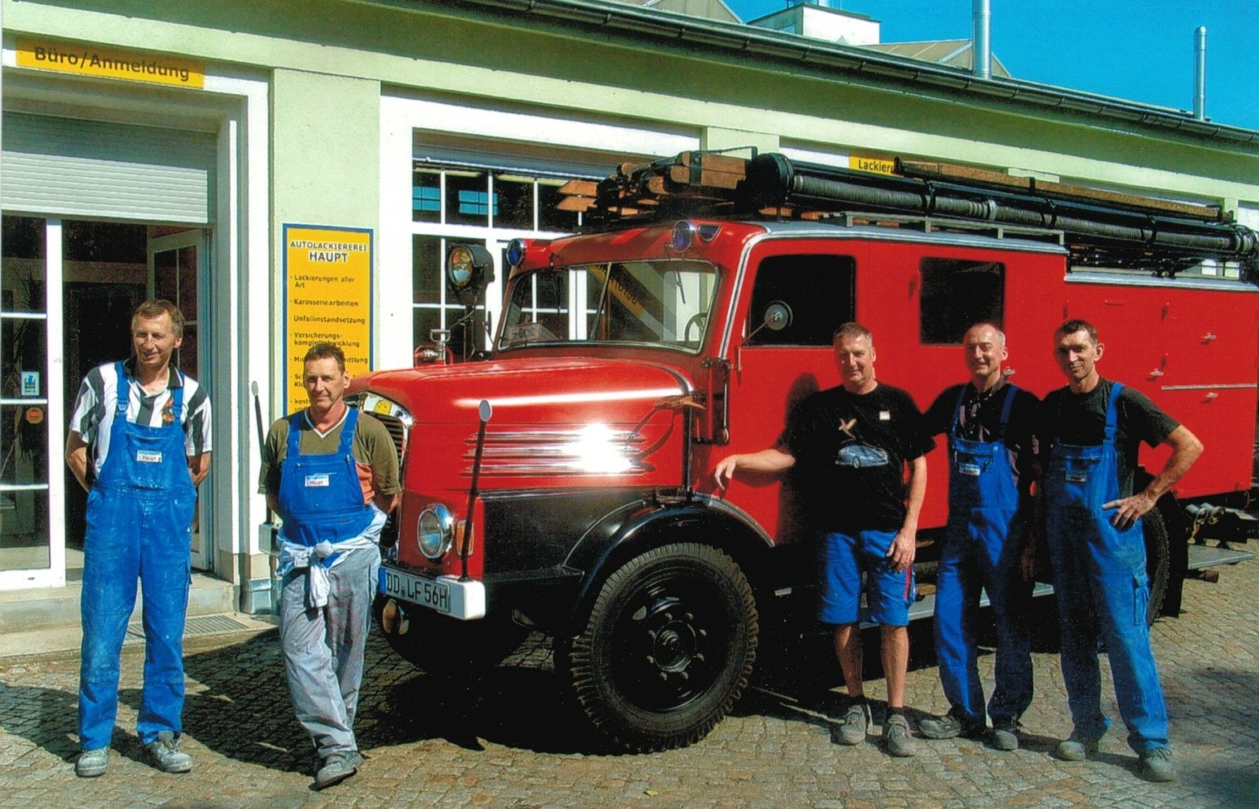 Vier Feuerwehrleute stehen neben einem roten Feuerwehrauto vor einer Feuerwache.