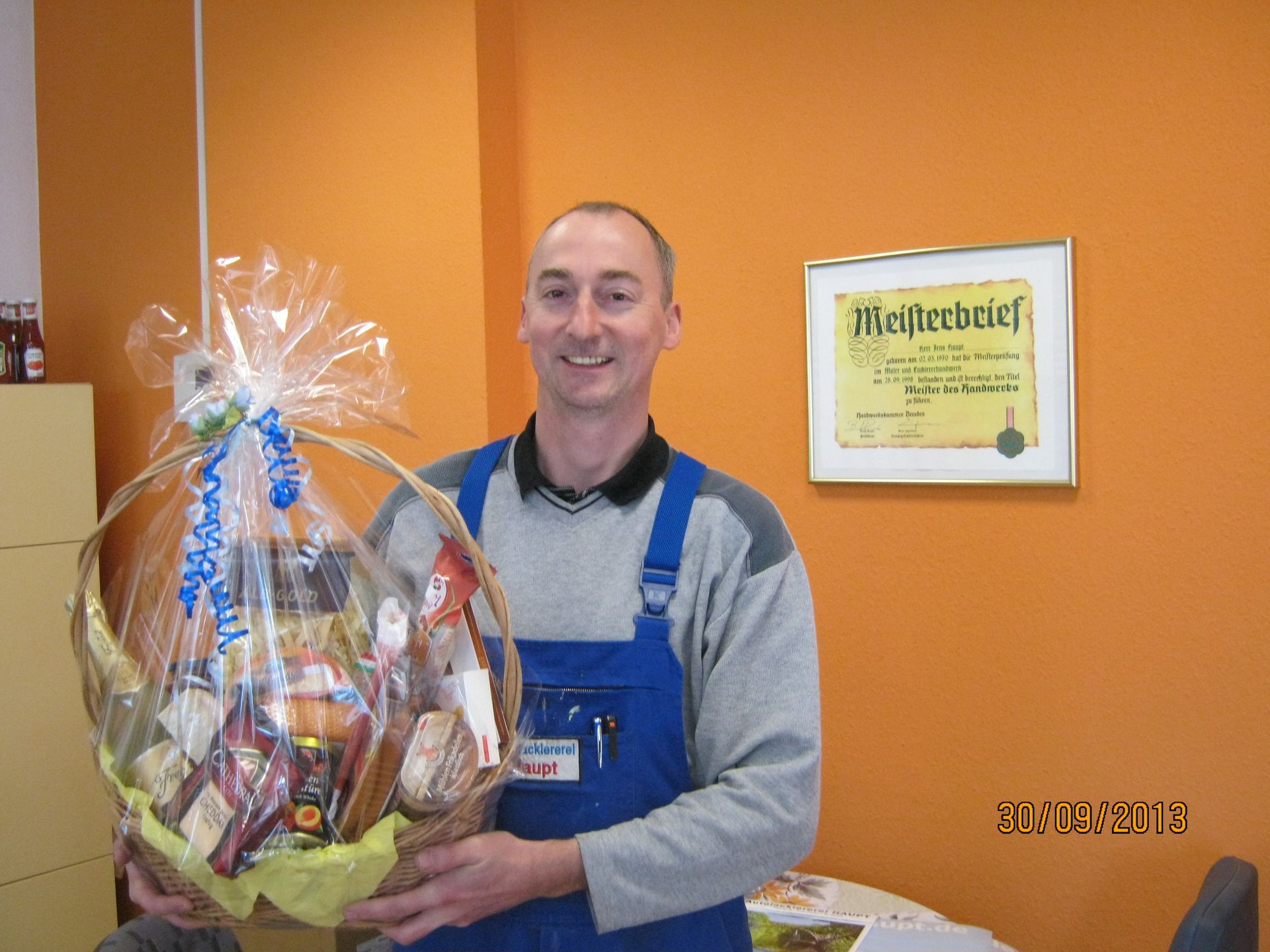 Ein Mann hält einen großen Geschenkkorb neben einer gerahmten Urkunde an einer orangefarbenen Wand.