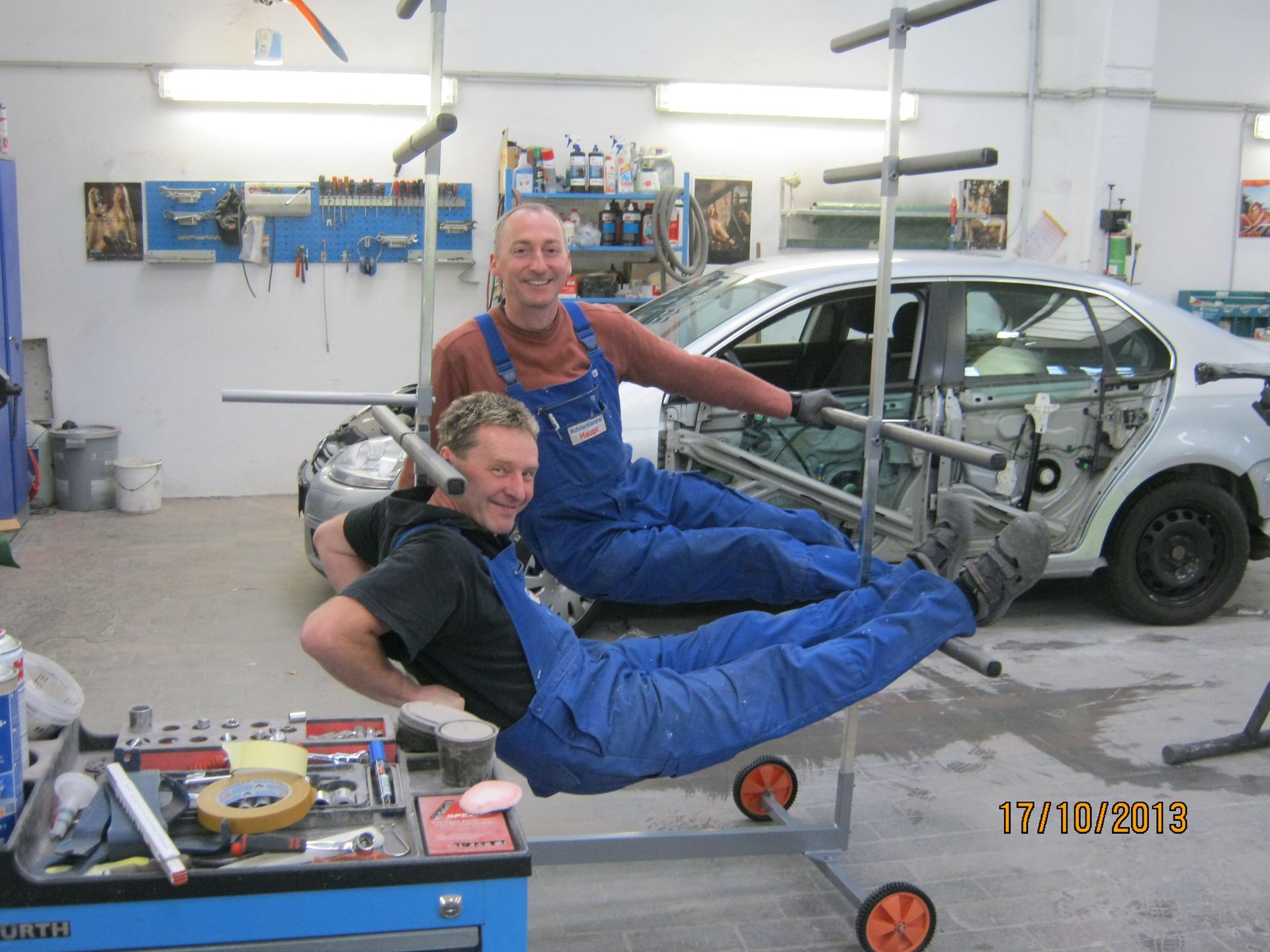 Zwei Mechaniker in blauen Overalls entspannen sich auf einem schwebenden Autochassis in einer Werkstatt.
