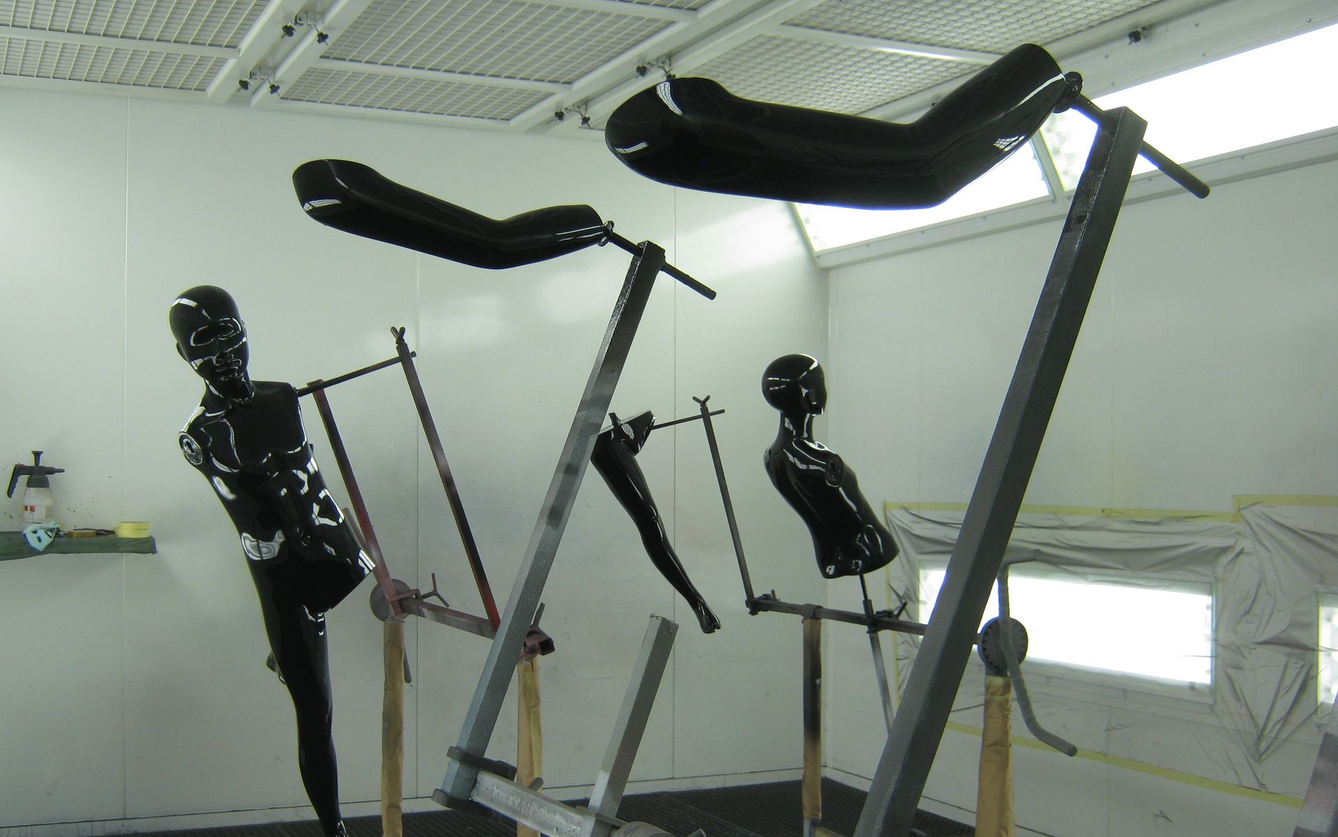 In einer weißen Werkstatt stehen zerlegte schwarze Fitnessgerätegestelle, deren Teile an Wänden und Boden lehnen.
