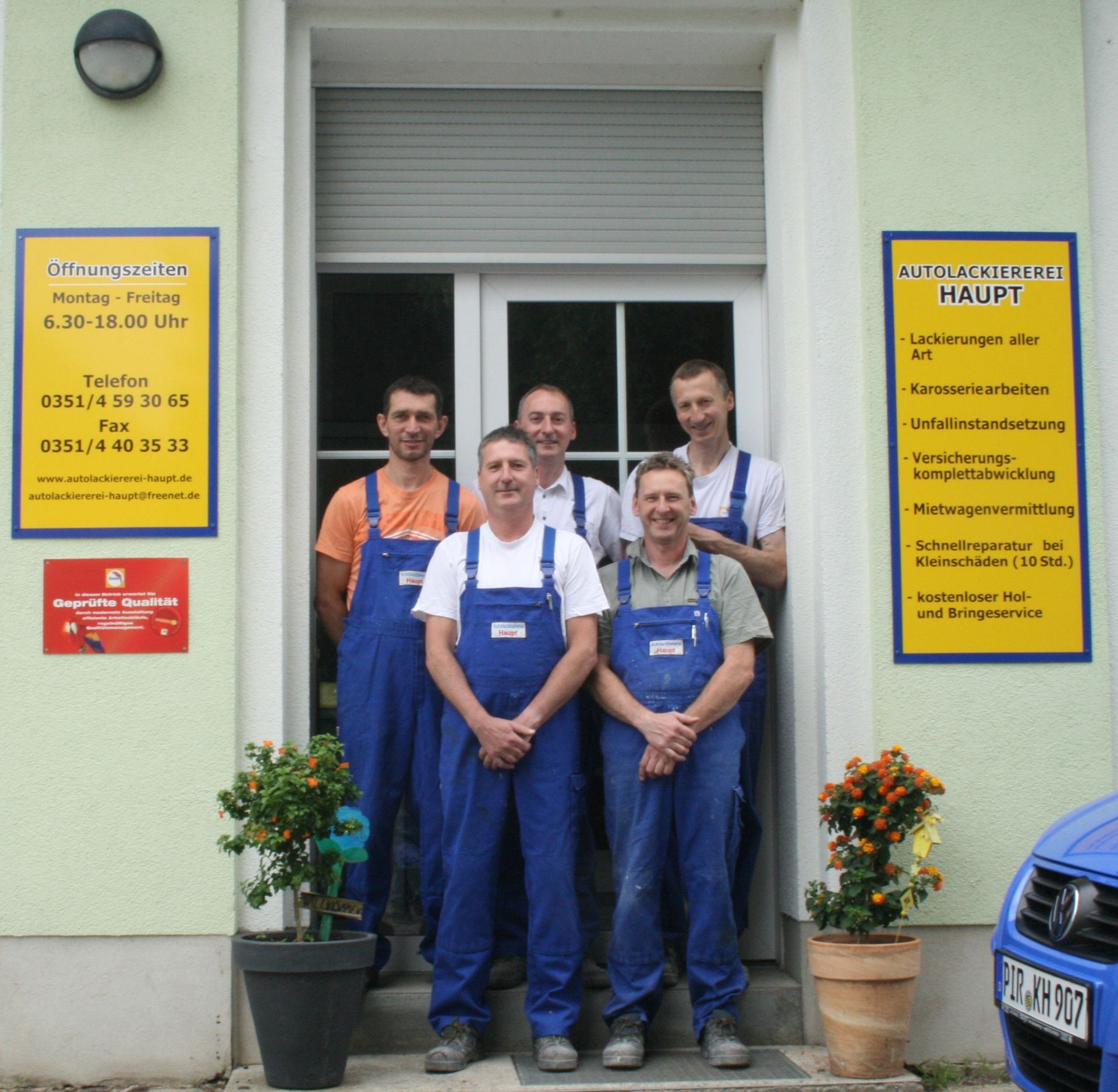 Fünf Arbeiter in blauen Overalls stehen am Eingang eines Geschäfts, an dessen Seiten sich gelbe Schilder befinden.