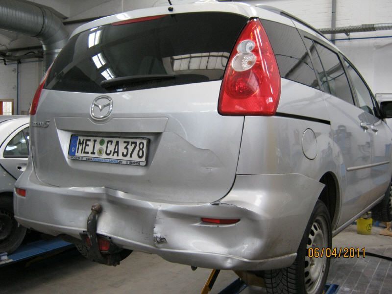 Silberner Mazda-Minivan mit Heckschaden in einer Garage, Kennzeichen sichtbar.