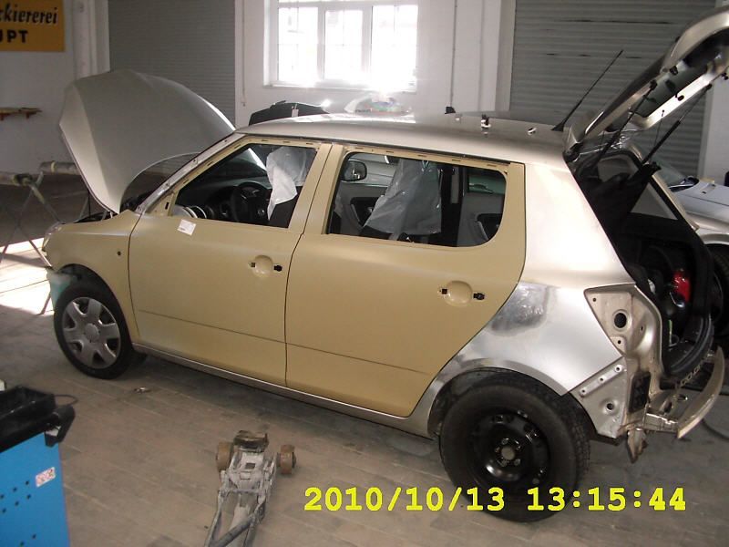 Beschädigter beiger Schrägheckwagen in einer Garage, Motorhaube geöffnet, Heck nach einem Zusammenstoß sichtbar.