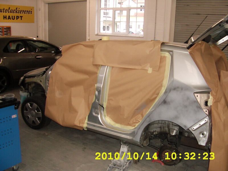 Die Karosserie eines Autos steht in einer Garage, mit Abdeckpapier und Klebeband für die Neulackierung abgedeckt; daneben steht ein weiteres Auto.