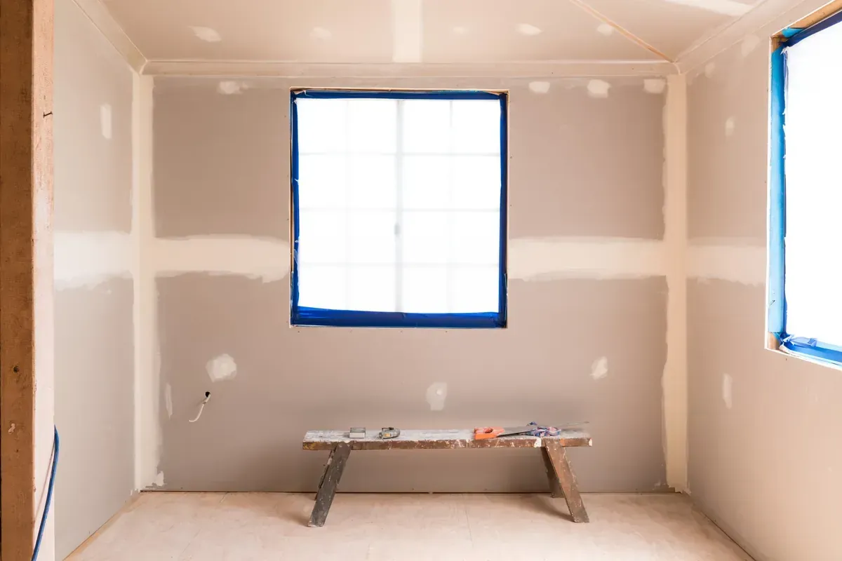 Habitación en construcción con paneles de yeso y ventanas selladas; un banco de trabajo.