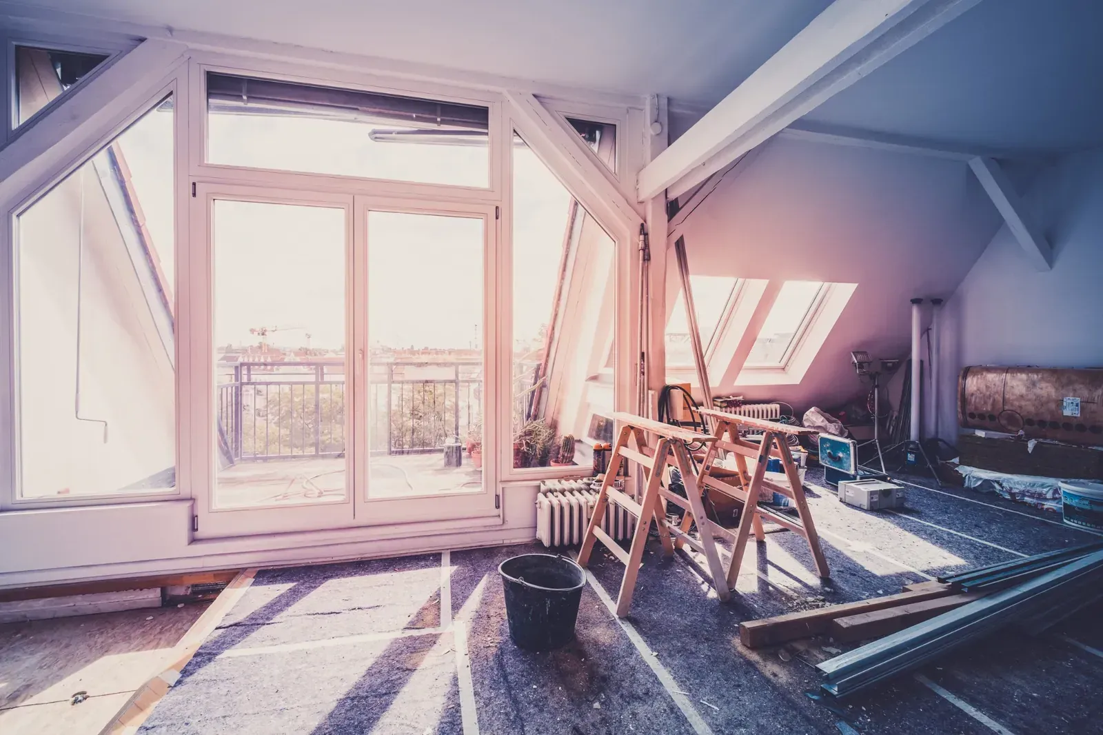 Interior de ático en reforma, con grandes ventanales, vigas de madera, escalera y restos de obra en el suelo.