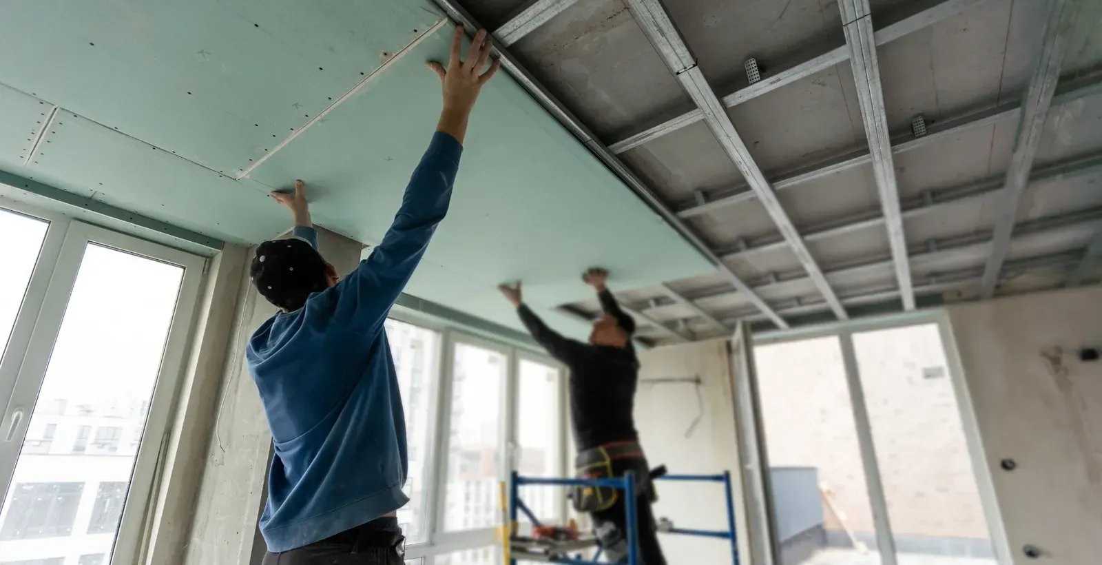 Los trabajadores instalan paneles de yeso en el marco del techo de una habitación con ventanas.