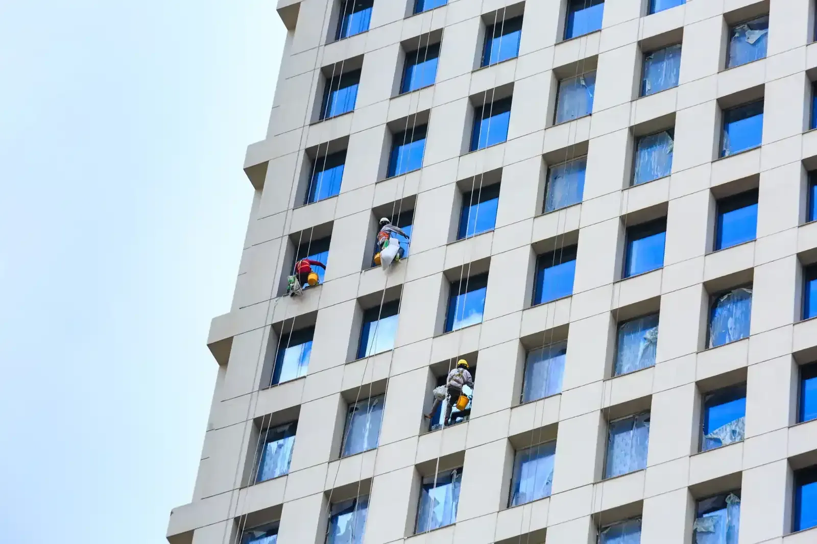 Limpiadores de ventanas con cuerdas limpiando un edificio alto con muchas ventanas bajo un cielo nublado.
