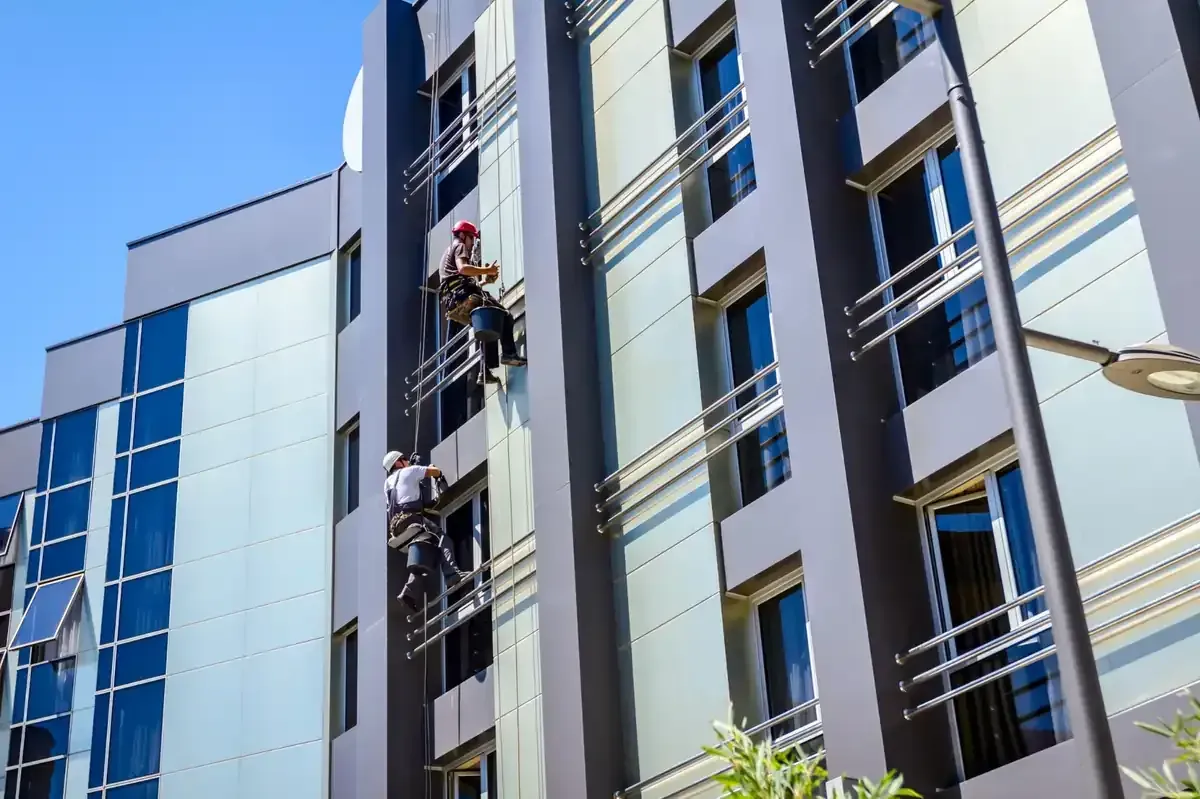 Dos limpiadores de ventanas descienden en rápel por el costado de un edificio alto y moderno bajo un cielo azul claro.