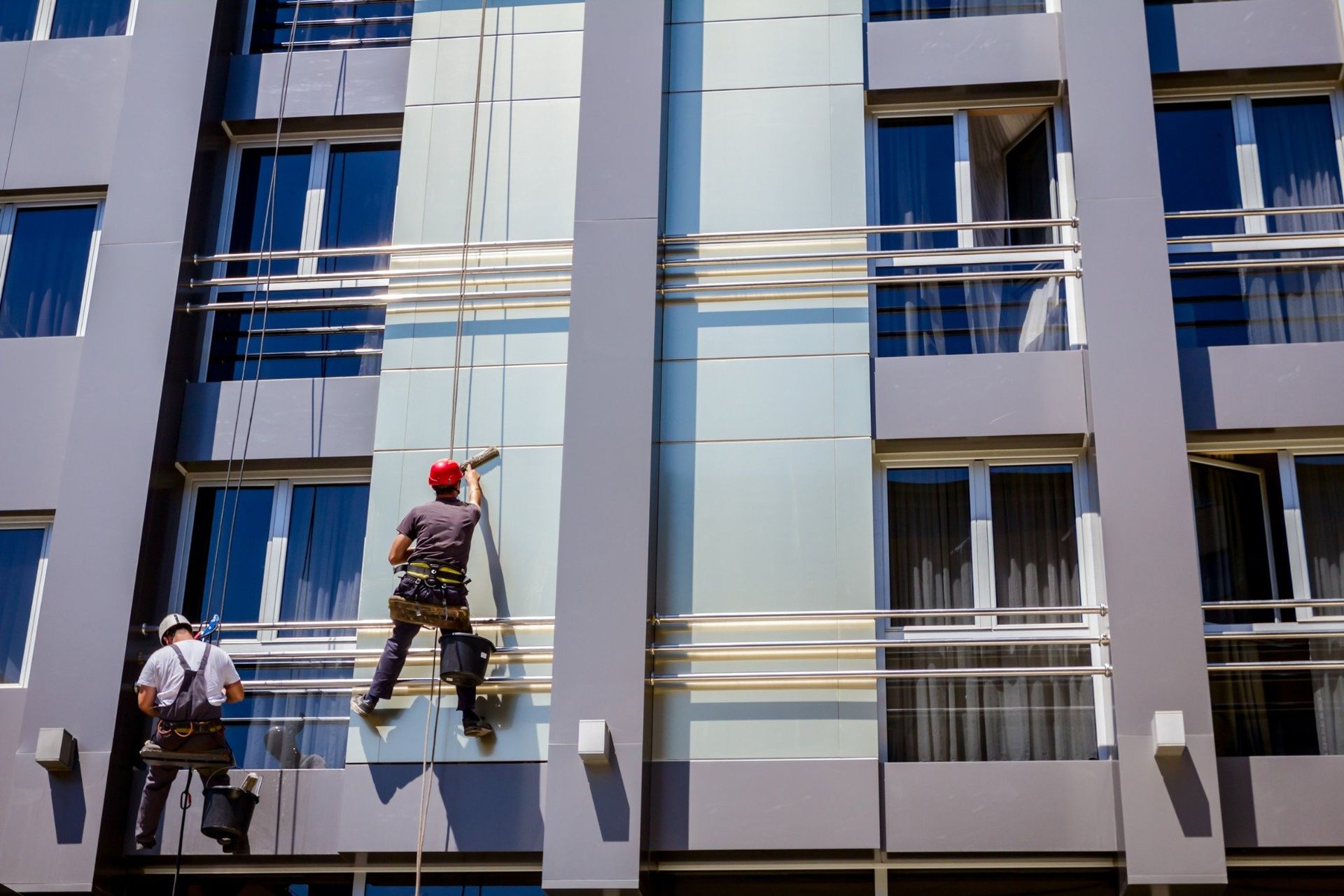 Limpiadores de ventanas limpiando paneles de vidrio en un edificio moderno, suspendidos por cuerdas.