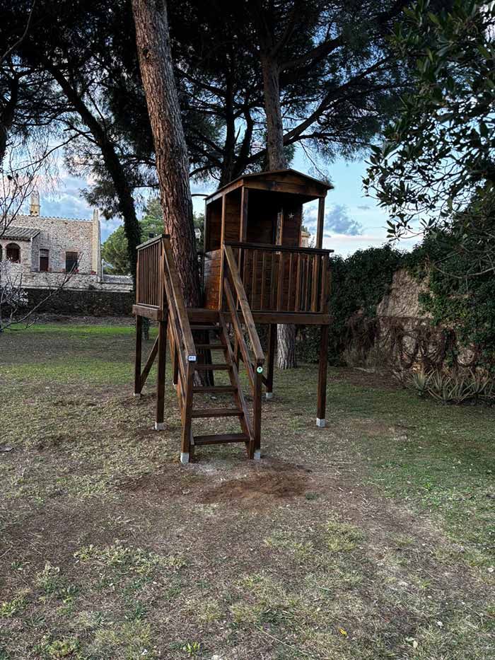 Una casa en el árbol de madera marrón con una escalera se encuentra sobre pilotes en un patio trasero cubierto de césped, cerca de un muro de piedra y árboles.