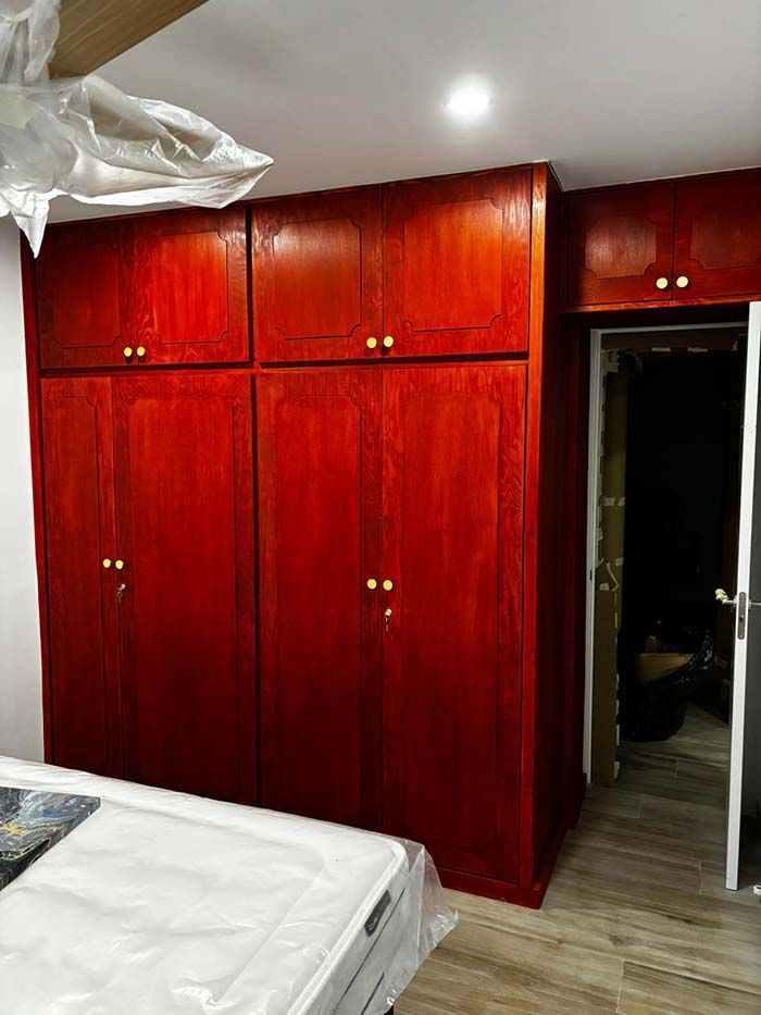 Un gran armario de madera de color rojo intenso con puertas dobles y gabinetes de almacenamiento superiores en un dormitorio al lado de una puerta abierta.