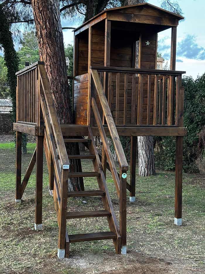 Una casa en el árbol de madera oscura construida sobre pilotes alrededor de un árbol, con una pequeña cabaña y una escalera de madera en un patio con césped.
